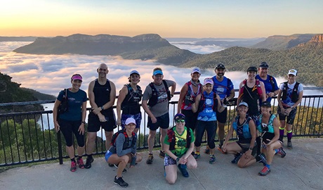 A group of runners stand on a lookout platform in front of a sweeping view of mountains and mist-filled valleys. Photo credit: Tony Williams © Blue Mountains Fitness